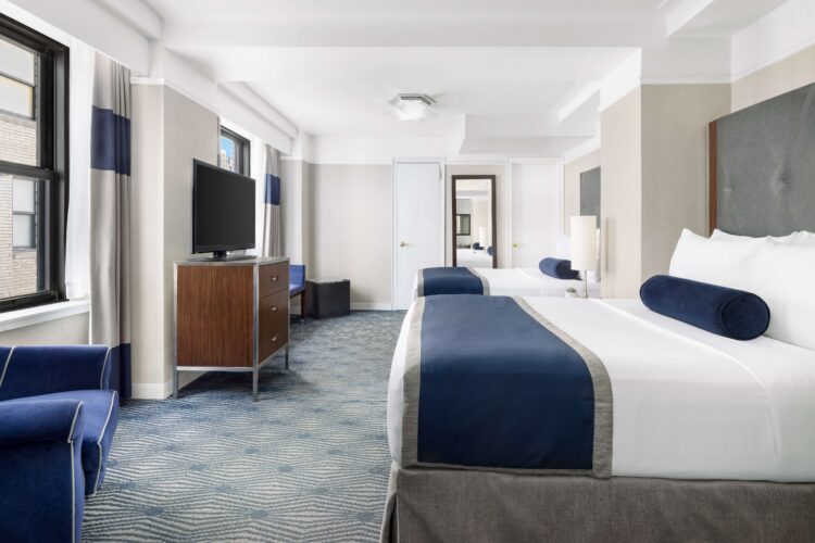 Spacious double room with two beds, desk area, armchair, and large TV in modern hotel setting