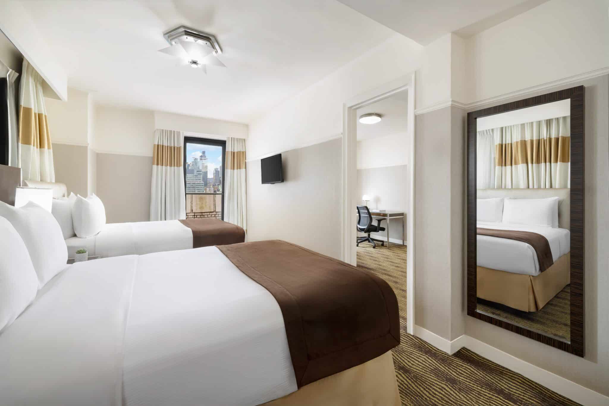 Family suite bedroom with two queen beds, wall mirror, TV, and adjoining workspace area
