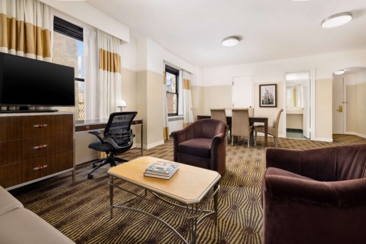Bright hotel suite with lounge seating, desk area, and large city-facing windows