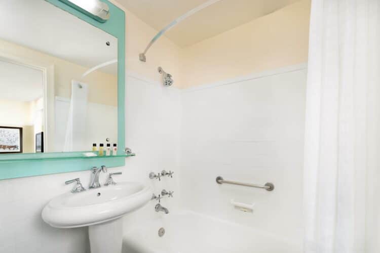 Hotel bathroom with bathtub and shower, white tiles, grab bar, and vanity with turquoise-framed mirror